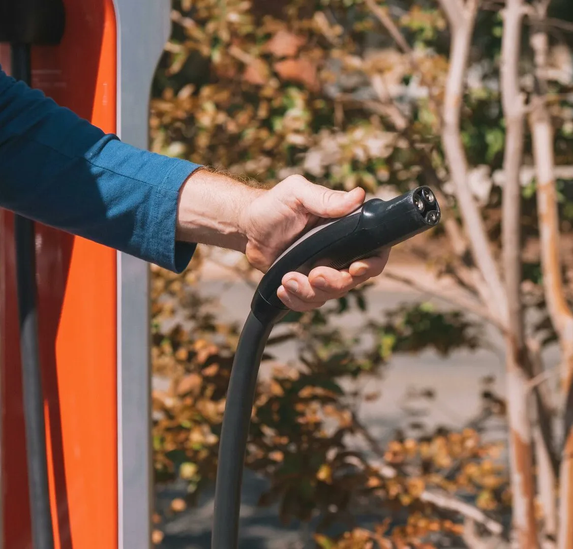Hand holding an electric vehicle charger at a station outdoors with autumn trees.