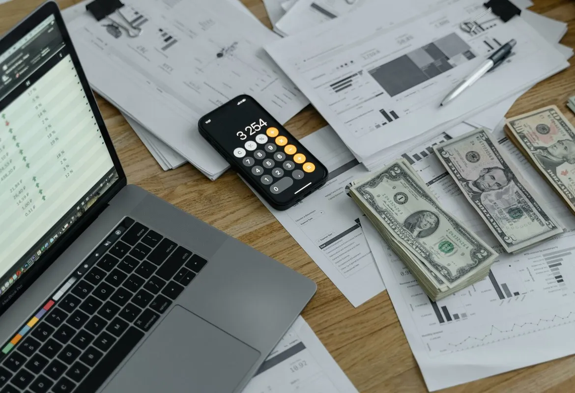 A desk with financial documents, currency, a laptop, and phone calculator.