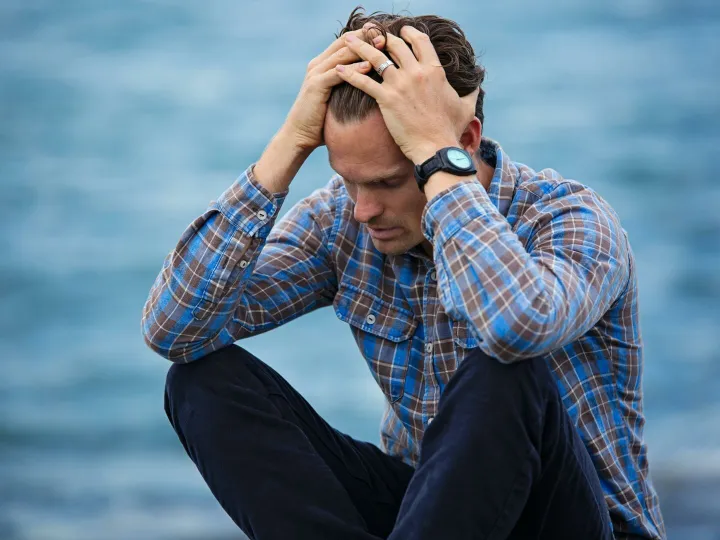 A man in a plaid shirt sits by the water looking distressed, symbolizing stress.