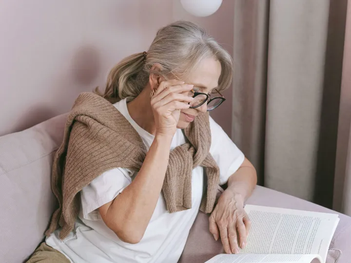 Elderly woman with glasses reading on a couch for leisure at home.