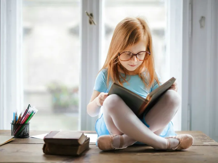 Adorable young girl wearing glasses, reading a book indoors by a window. Captures innocence and focus.