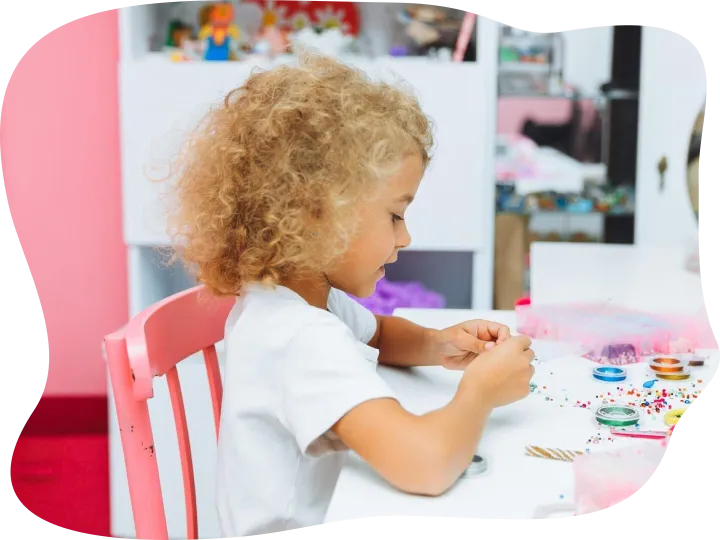 Cute little blonde girl making bead jewelry at a table in th
