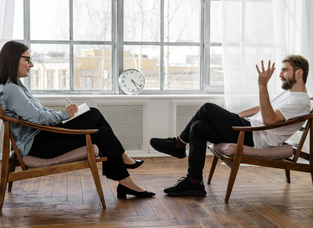 Person in Black Pants and Black Shoes Sitting on Brown Wooden Chair