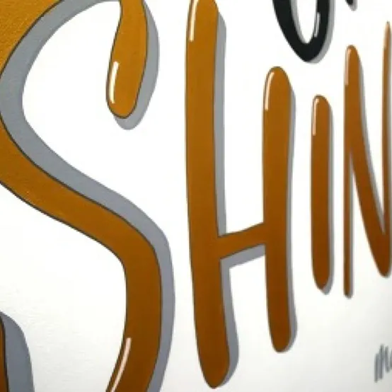 The word Shine hand lettered in gold on white board