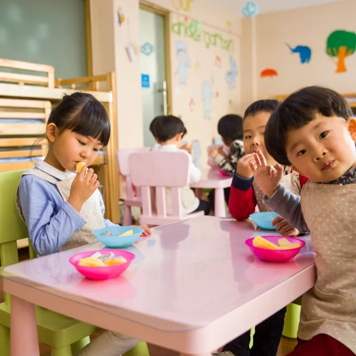 Young children sitting at a small table in a nursery eating fruit at snack time