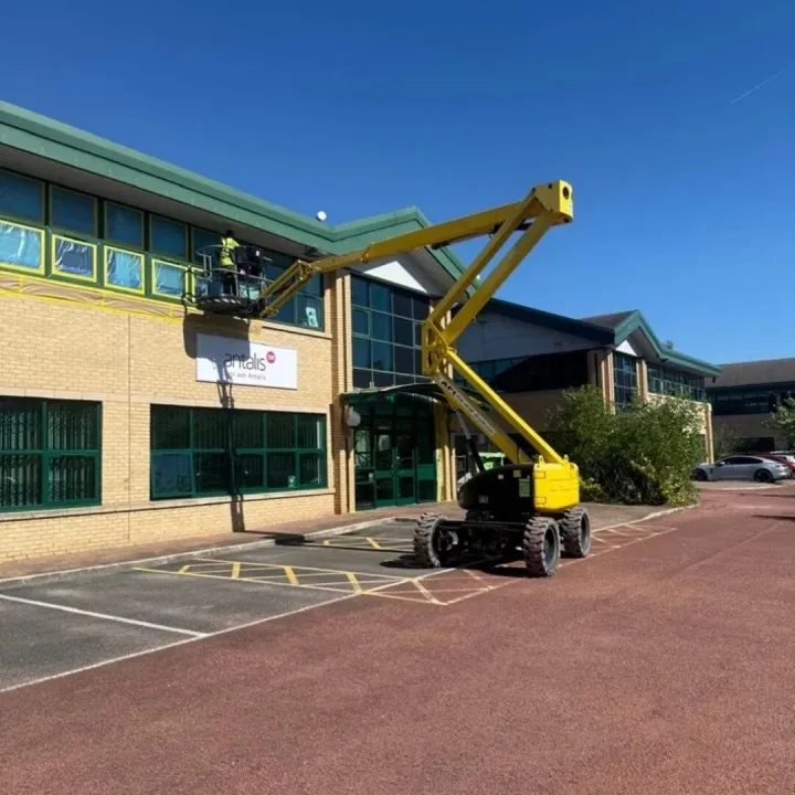 Operative in a boom lift working on masked green window frames of a brick office building