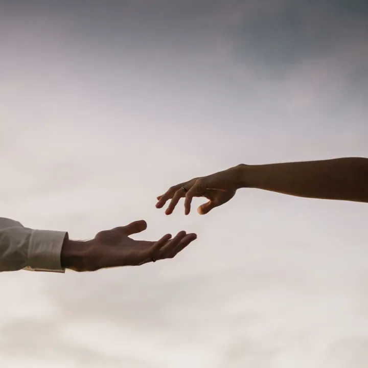 Two hands reaching towards each other against a soft twilight sky symbolizing connection and love.