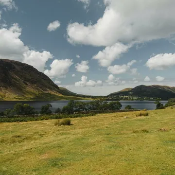 Grassy meadow located near calm river and green hills against cloudy blue sky on sunny day in nature