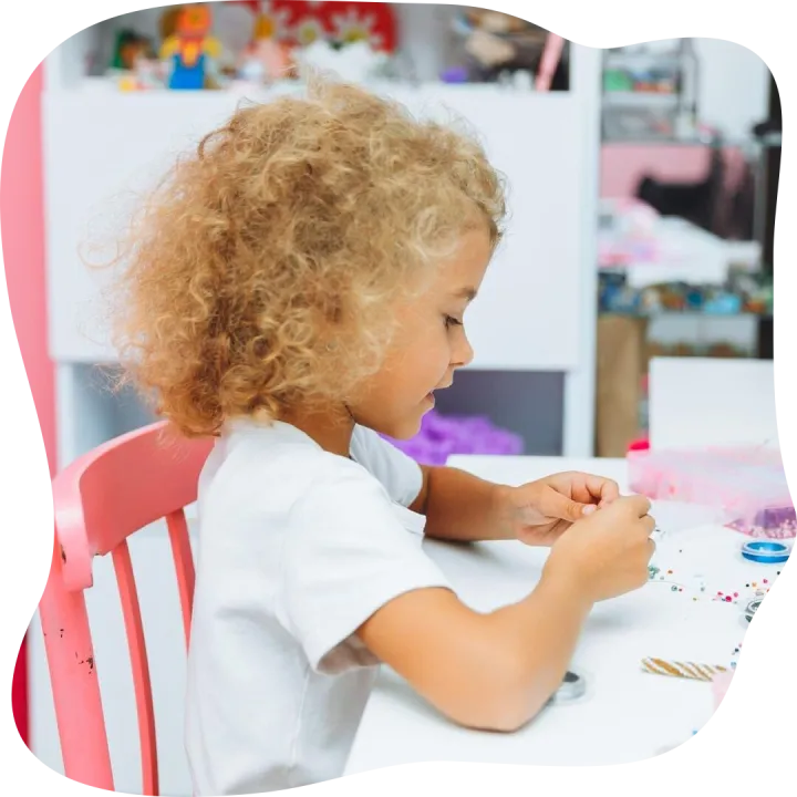 Cute little blonde girl making bead jewelry at a table in th
