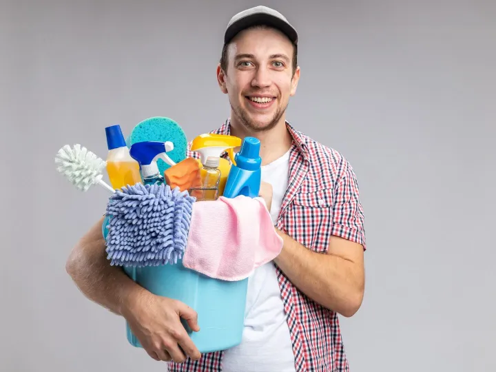 Smiling cleaner in a cap holding a bucket of cleaning supplies