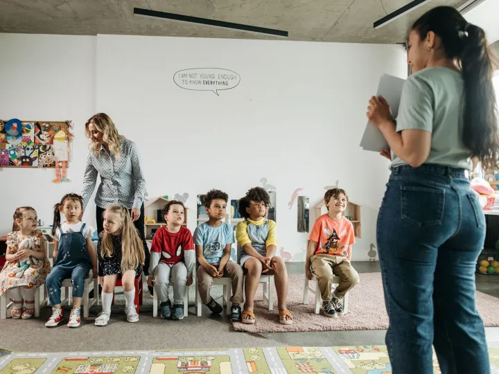 A group of preschool children sitting on small chairs with two practitioners in a nursery room