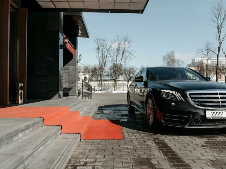 A luxury black sedan parked on a red carpet driveway outside a modern building.
