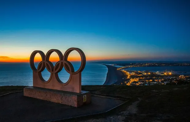 Scenic sunset view of the Olympic rings monument by the sea with city lights in the distance.