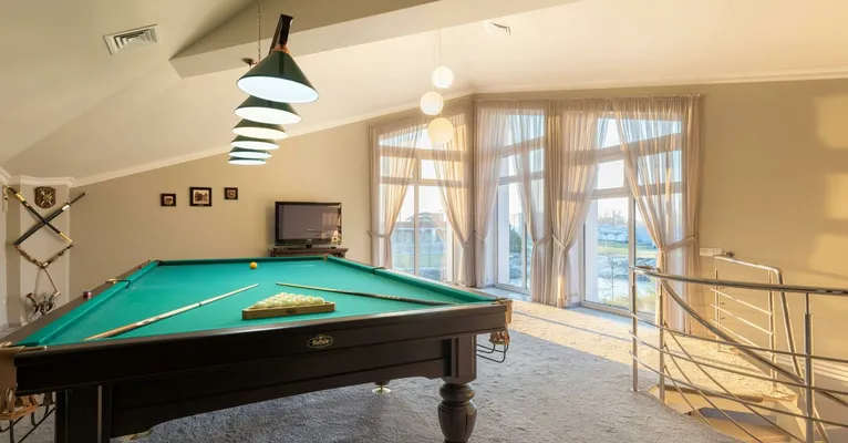 Wooden pool table with cues placed on carpet in spacious room with windows and staircase with metal railing in contemporary building