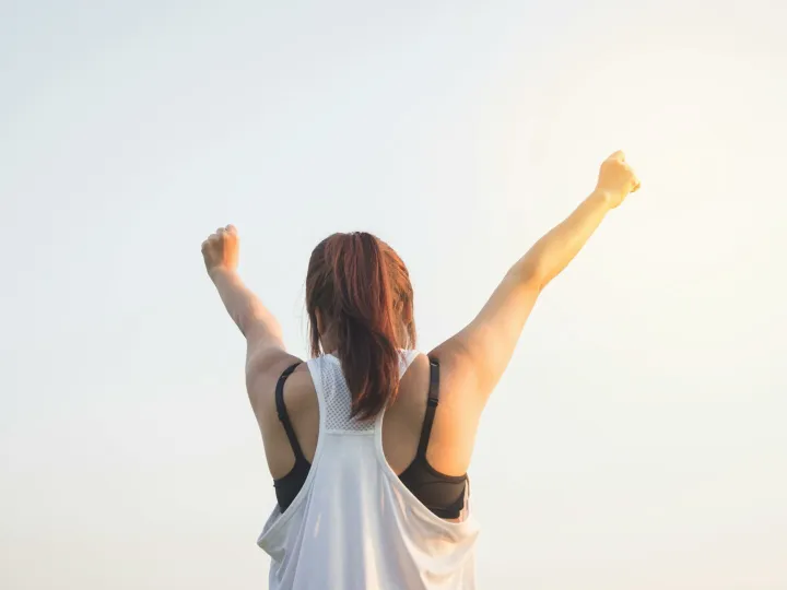 A woman celebrates success with arms raised in a bright outdoor setting.